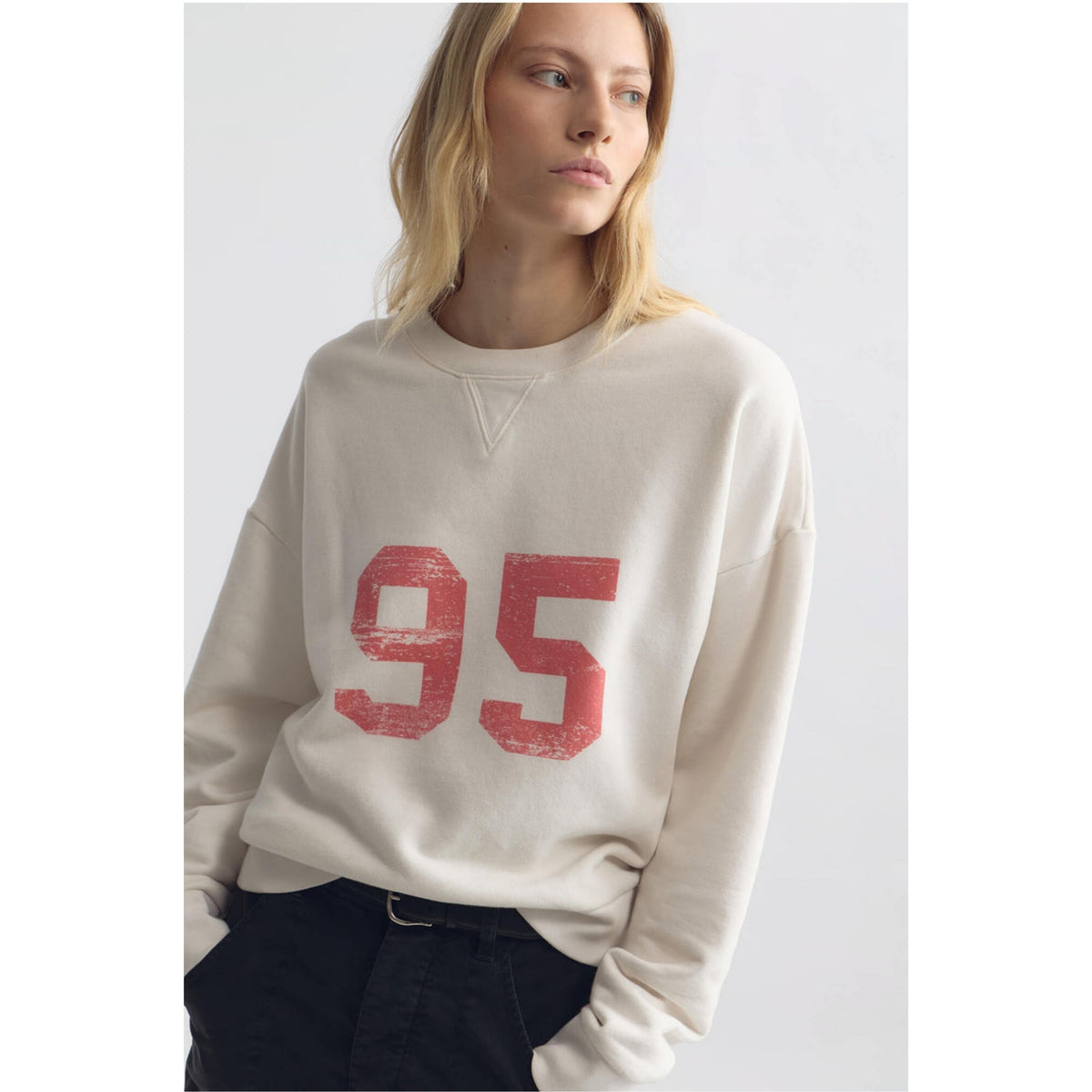 A woman wearing a cream-colored crewneck sweatshirt with a distressed red '95' graphic on the front, paired with black pants, against a light gray studio background.