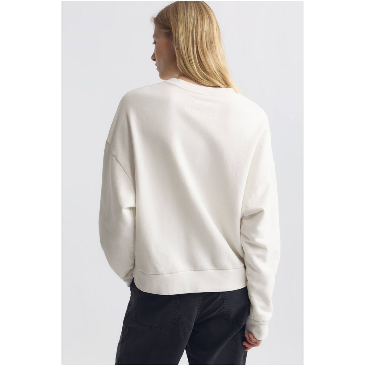 A woman viewed from the back wearing an oversized off-white crewneck sweatshirt and dark pants against a light gray studio background.