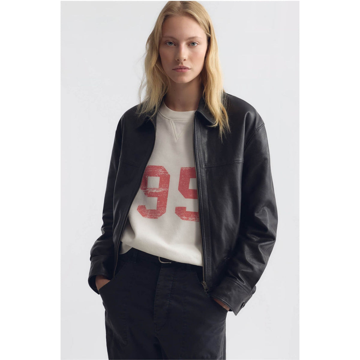 A blonde woman wearing a black leather jacket over a cream crewneck sweatshirt with a distressed red '99' graphic, paired with black trousers, against a light gray studio background.