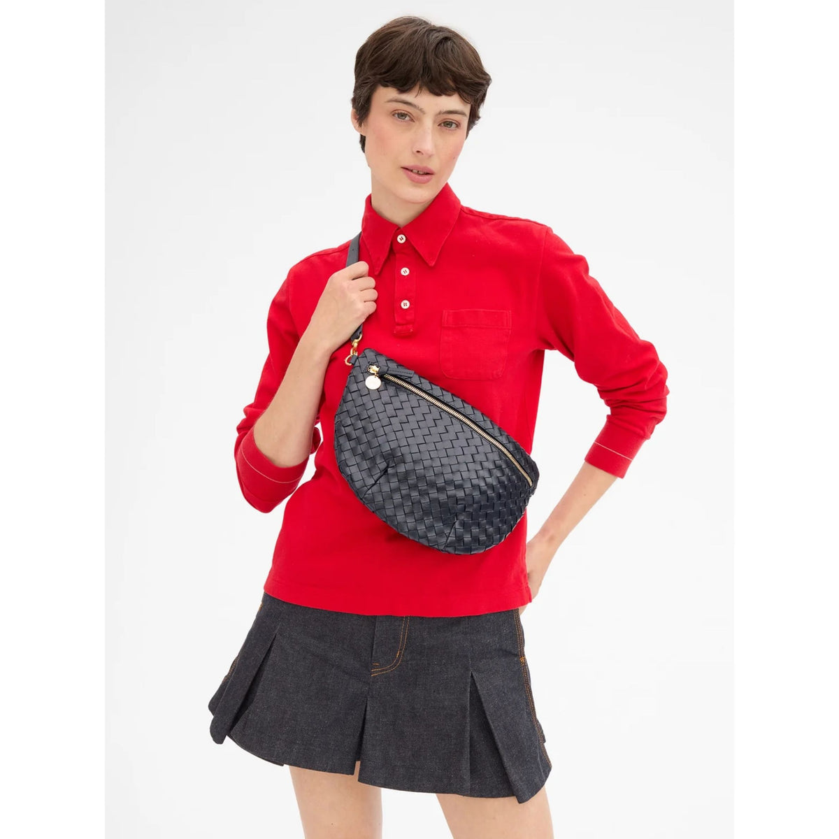 A model wearing a red long-sleeve polo shirt and a dark gray pleated mini skirt, carrying a dark gray woven patterned crossbody bag with a silver zipper on a plain white background.