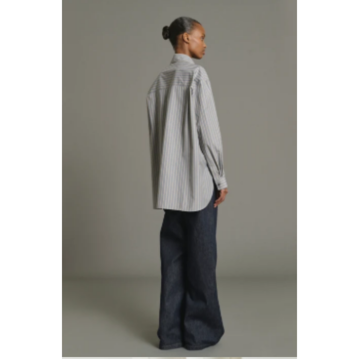 A model shown from the back wearing a light gray and white vertically striped button-down shirt and dark wide-leg jeans, standing against a solid gray studio background.