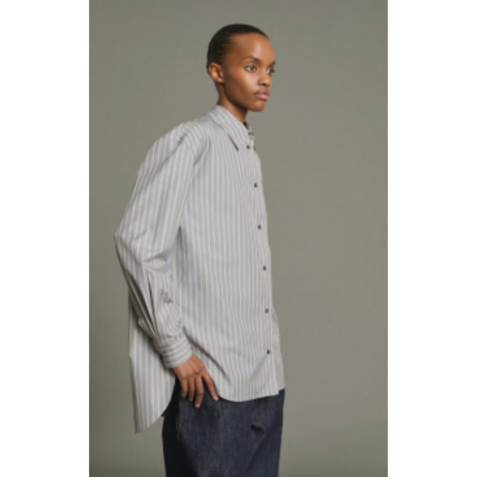 A model wearing a light gray and white vertically striped long-sleeve button-down shirt with dark blue jeans, standing against a solid gray studio background.