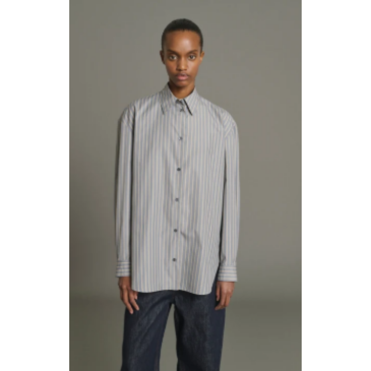 A model wearing a light gray and white vertically striped long-sleeve button-down shirt with dark blue jeans, standing against a solid gray studio background.