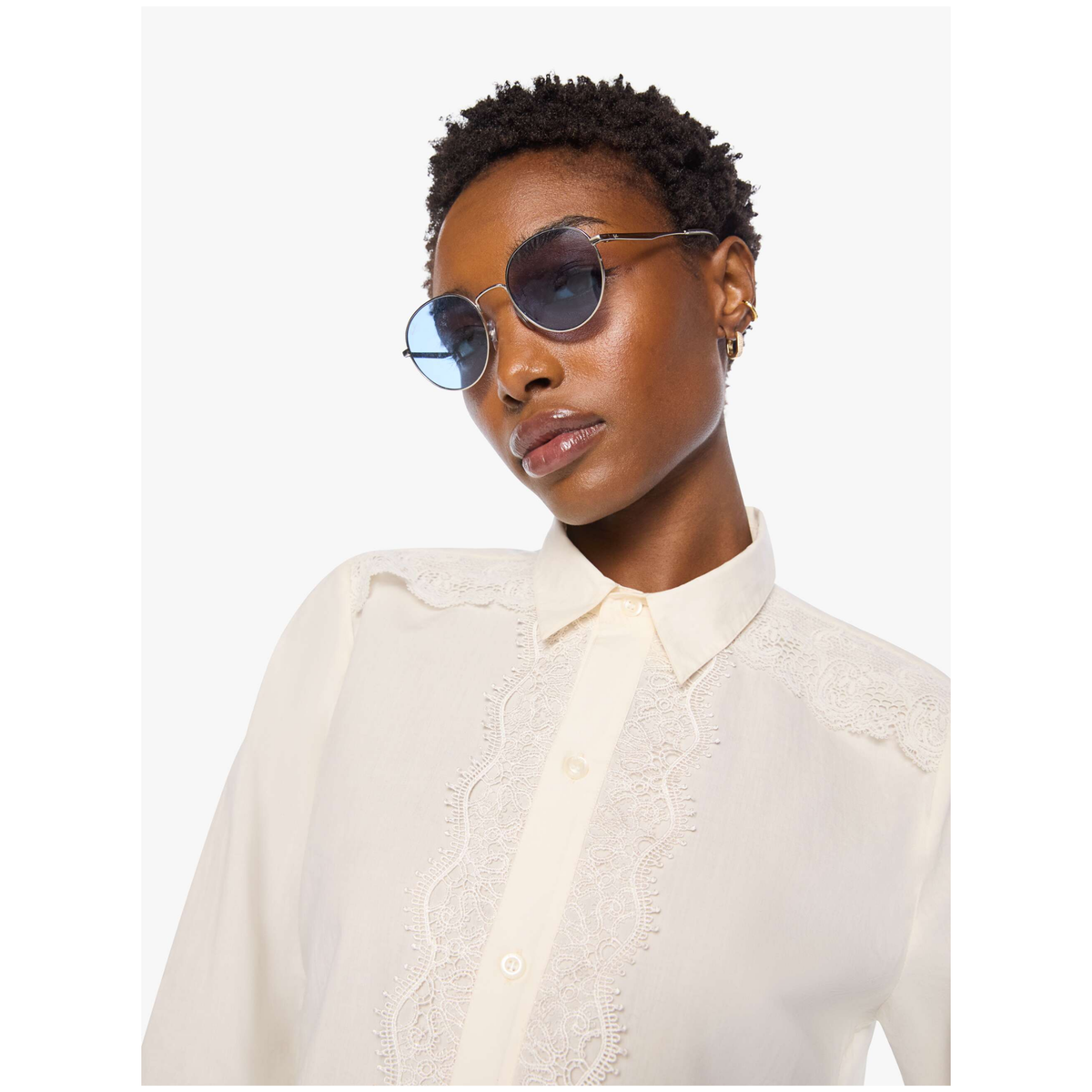 A model wearing round sunglasses with blue-tinted lenses and slim gold metal frames, paired with a cream-colored lace-trimmed blouse, set against a plain light gray background.
