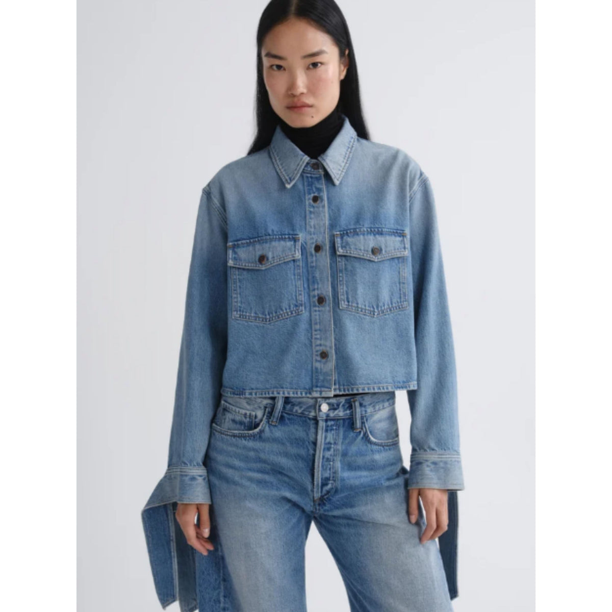 A model wearing a cropped light blue denim shirt jacket with chest flap pockets, paired with matching blue jeans and a black turtleneck, against a light gray studio background.