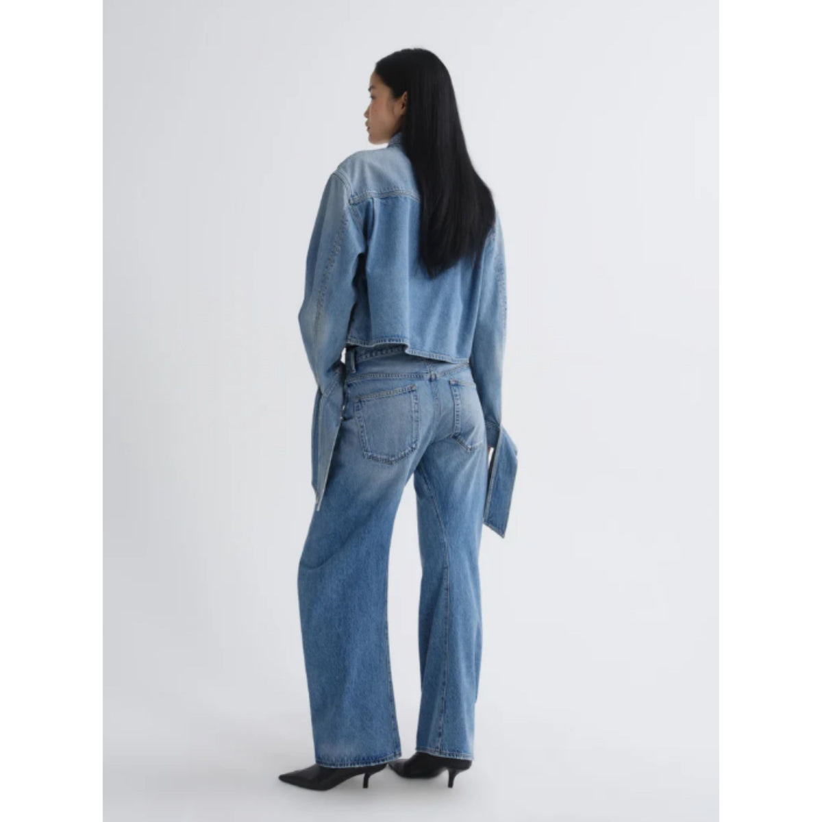 A model is shown from the back wearing a light blue cropped denim jacket and matching wide-leg denim jeans, paired with black pointed-toe heels against a plain light gray studio background.