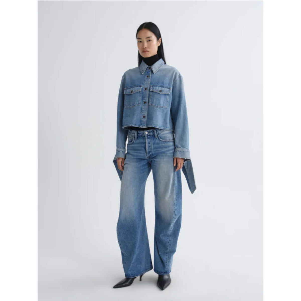 A model stands against a light gray studio background wearing a cropped light-wash blue denim shirt-jacket with matching wide-leg jeans, a black turtleneck, and black pointed heels.