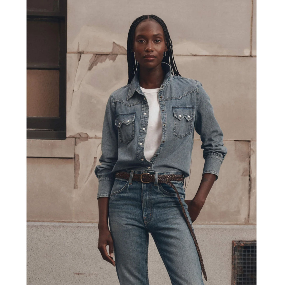 A model stands against a textured beige stone wall wearing a light-wash blue denim shirt jacket with silver studs, a white t-shirt, and blue jeans with a brown belt.