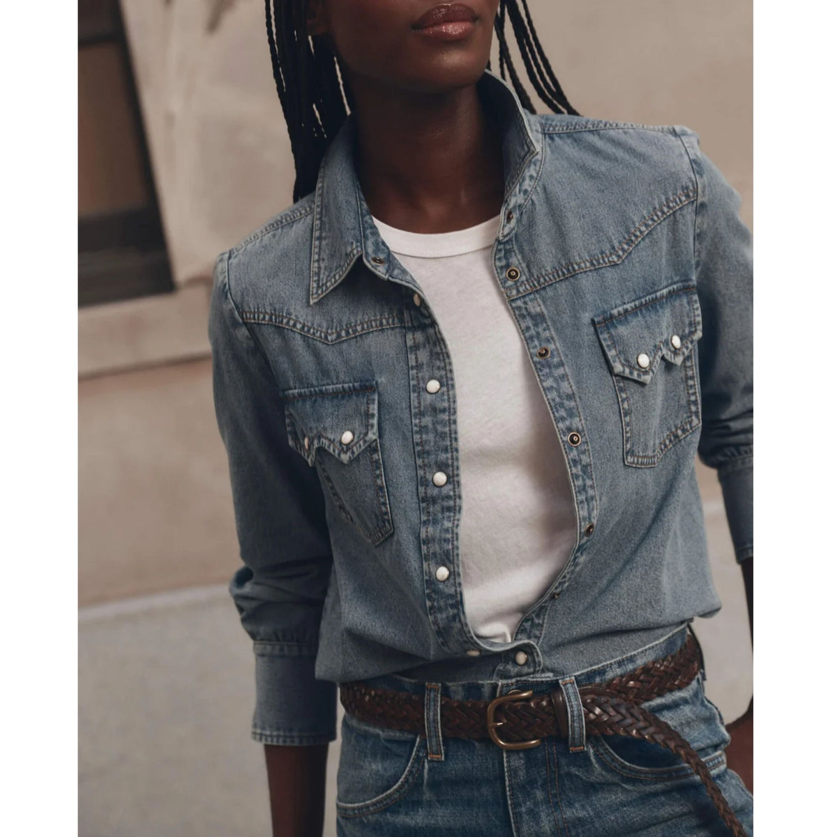 A woman wearing a light-wash blue denim western-style shirt jacket over a white t-shirt, paired with blue jeans and a brown braided belt.