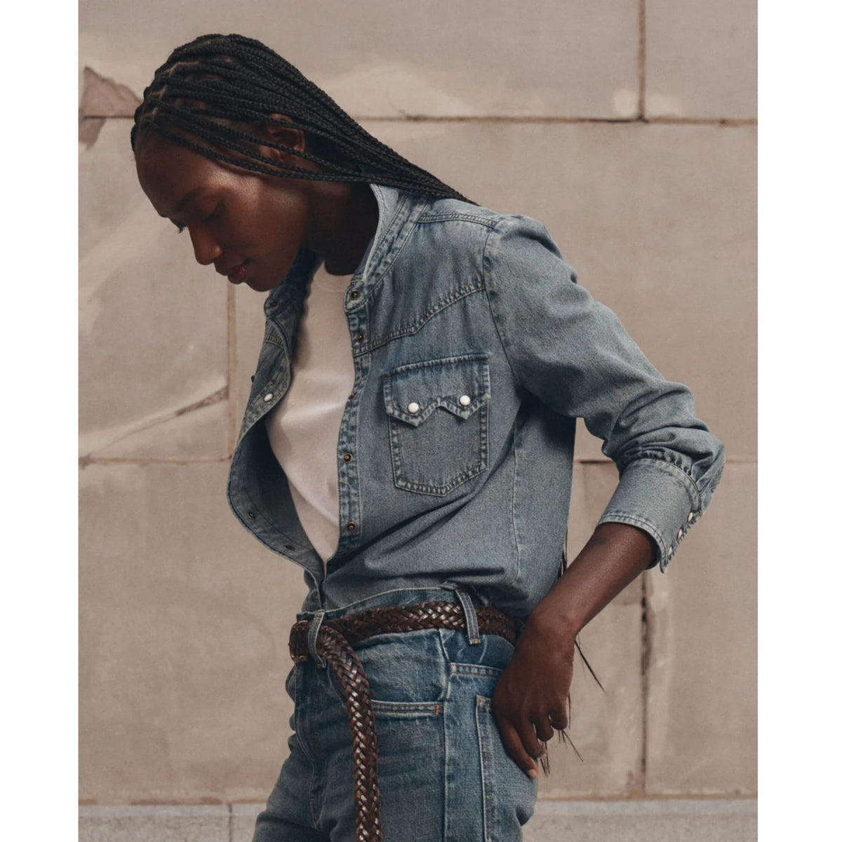 A woman wearing a light blue denim jacket over a white t-shirt and blue jeans, styled with a brown braided belt, standing against a beige stone wall.