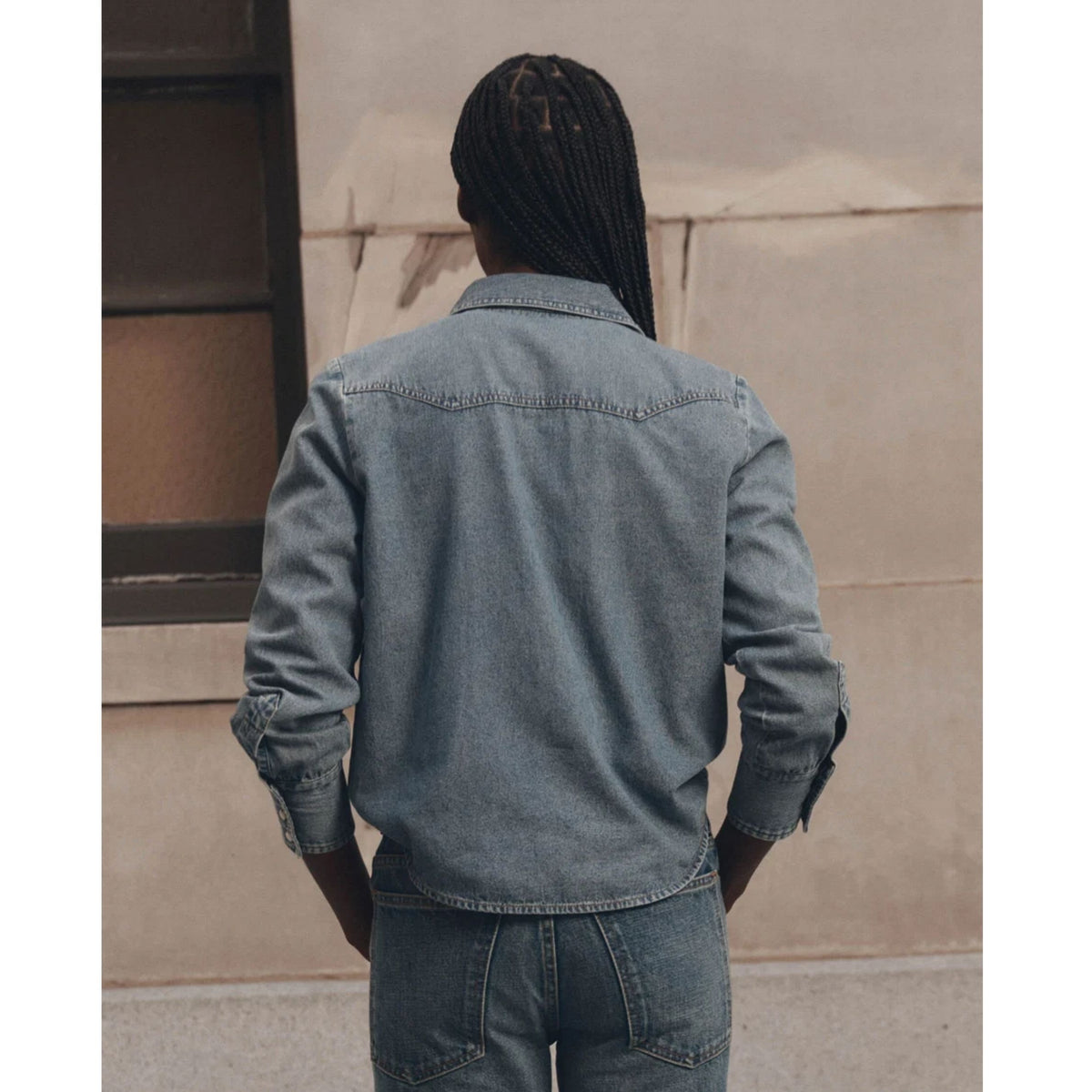 A person shown from behind wearing a light blue denim shirt jacket and matching blue jeans, standing against a beige wall.