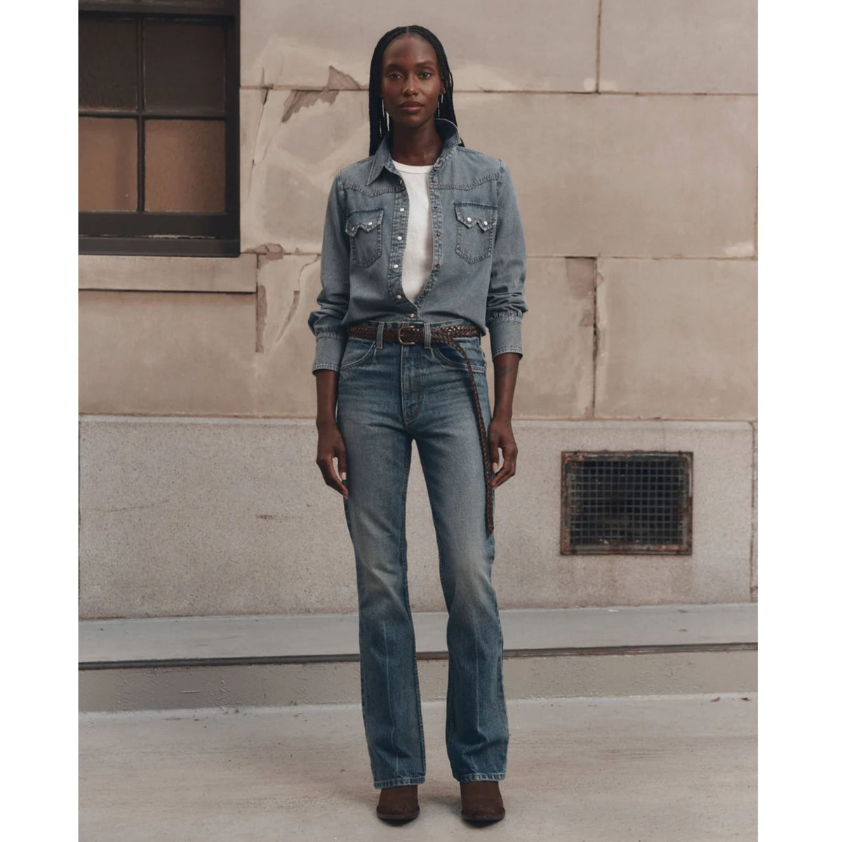 A model stands outdoors against a beige stone wall wearing a light-wash blue denim shirt-jacket and matching blue jeans with a brown belt.