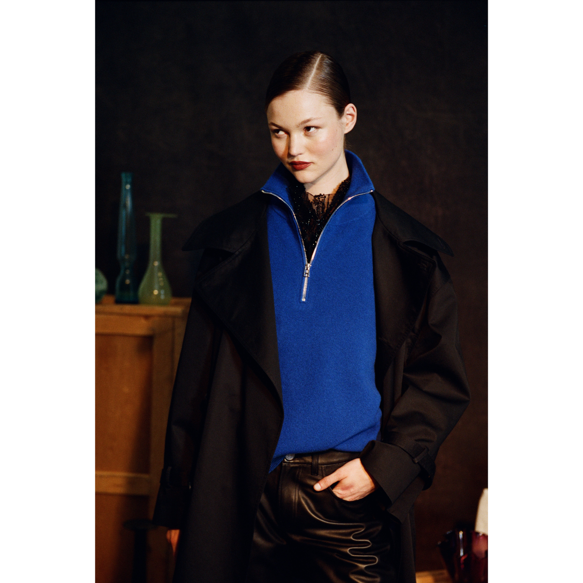 A model wearing a black coat over a bright blue quarter-zip sweater and black leather pants, posed against a dark, moody background with a wooden cabinet and green glass vases.
