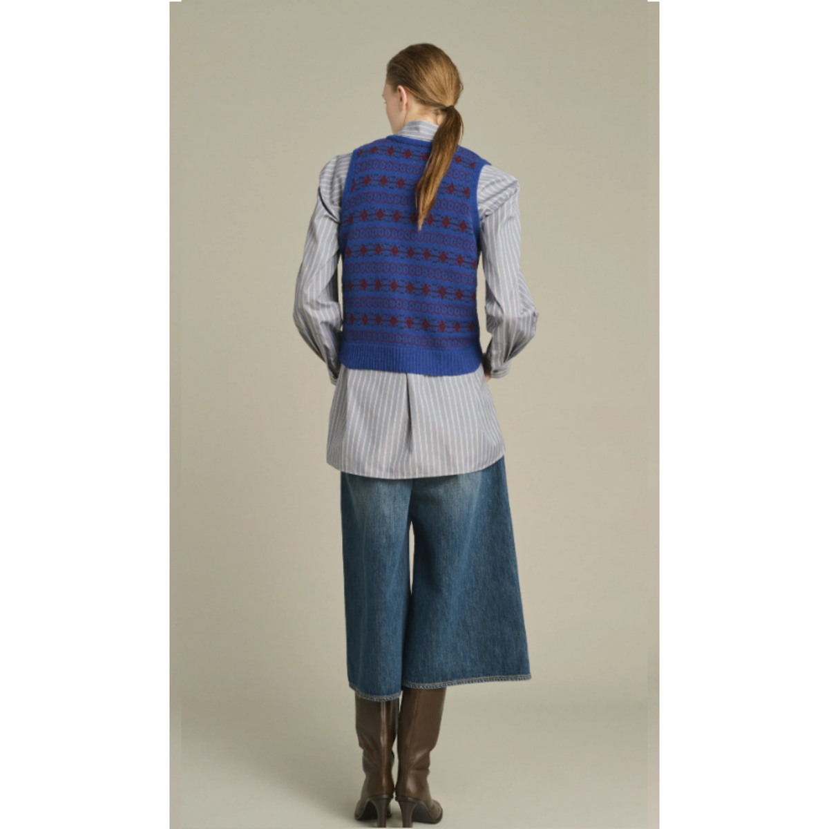 A woman shown from the back wearing a blue patterned vest over a light gray and white striped shirt, blue denim culottes, and brown knee-high boots, against a plain beige studio background.