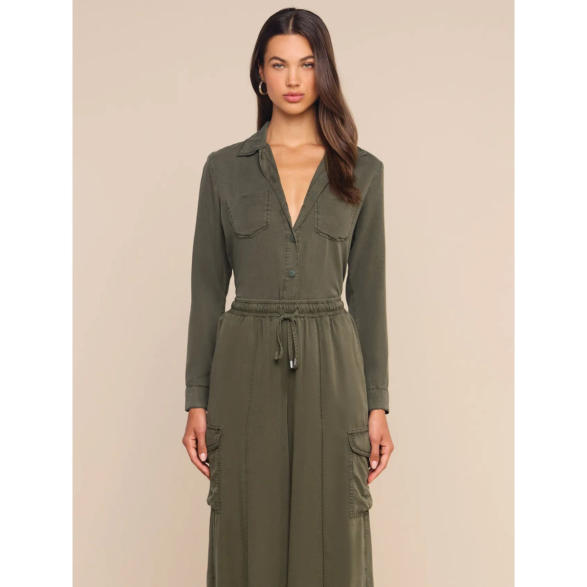 A woman modeling an olive green long-sleeve utility jumpsuit with a button-front chest, deep V-neck, drawstring waist, and cargo pockets, set against a solid beige background.