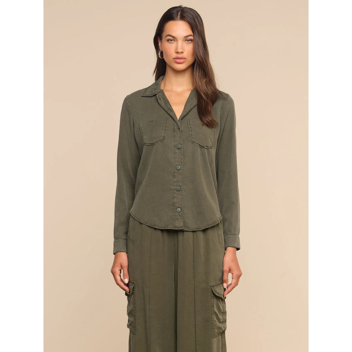 A woman modeling an olive green matching set consisting of a long-sleeve button-front shirt with chest pockets and matching cargo pants, set against a solid beige studio background.