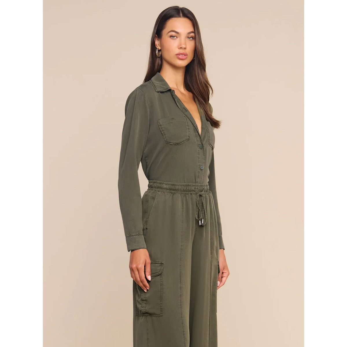 A woman modeling an olive green utility jumpsuit with a collar, button front, drawstring waist, and cargo pockets, set against a solid beige background.
