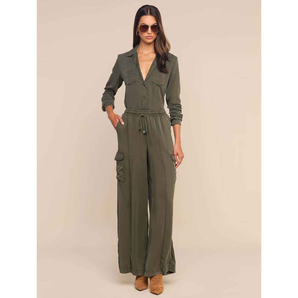 A woman modeling an olive green utility jumpsuit with a button-front bodice, long sleeves, side cargo pockets, and a drawstring waist, photographed against a plain beige studio background.