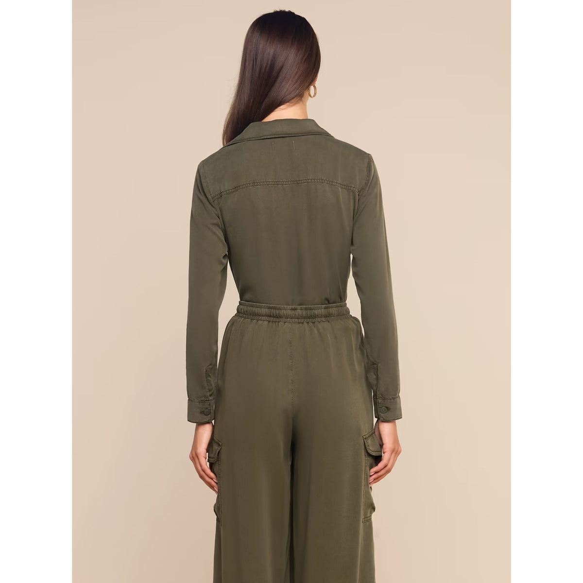 A woman viewed from the back wearing an olive green utility jumpsuit with a collar, long sleeves, and a drawstring waist, set against a solid beige background.
