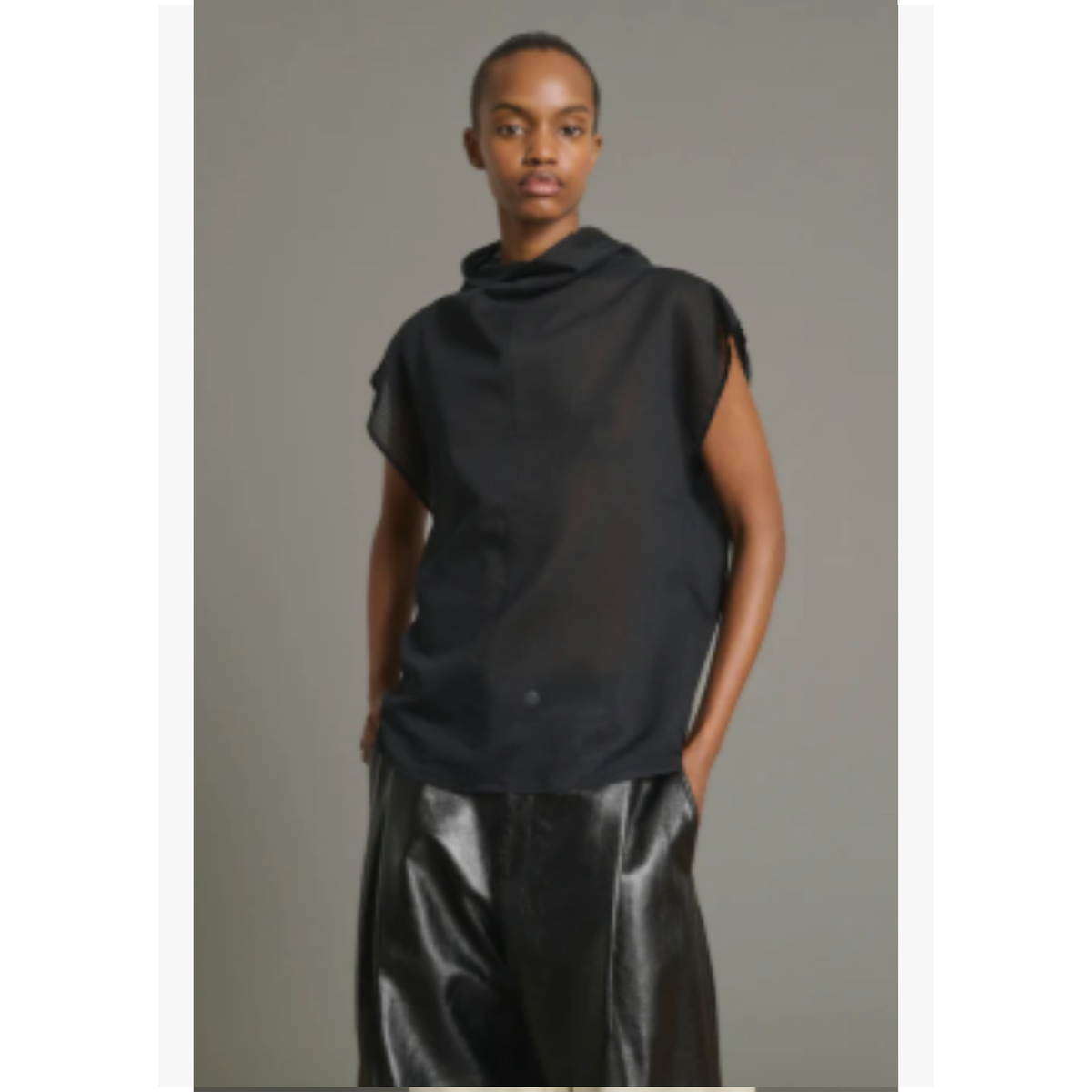 A model wearing a black sleeveless draped top and shiny black pants, standing against a plain gray studio background.