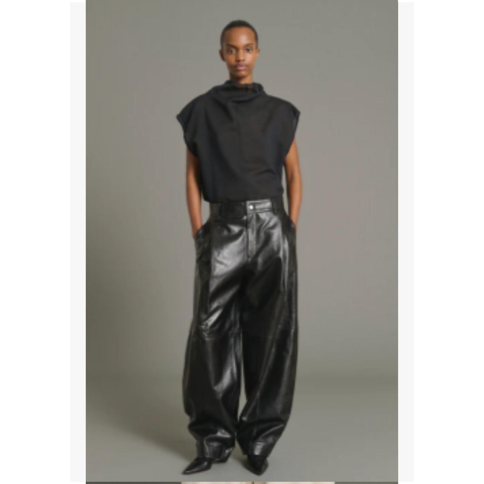 A model wearing a black sleeveless high-neck top and shiny black wide-leg trousers, standing against a neutral gray studio background.