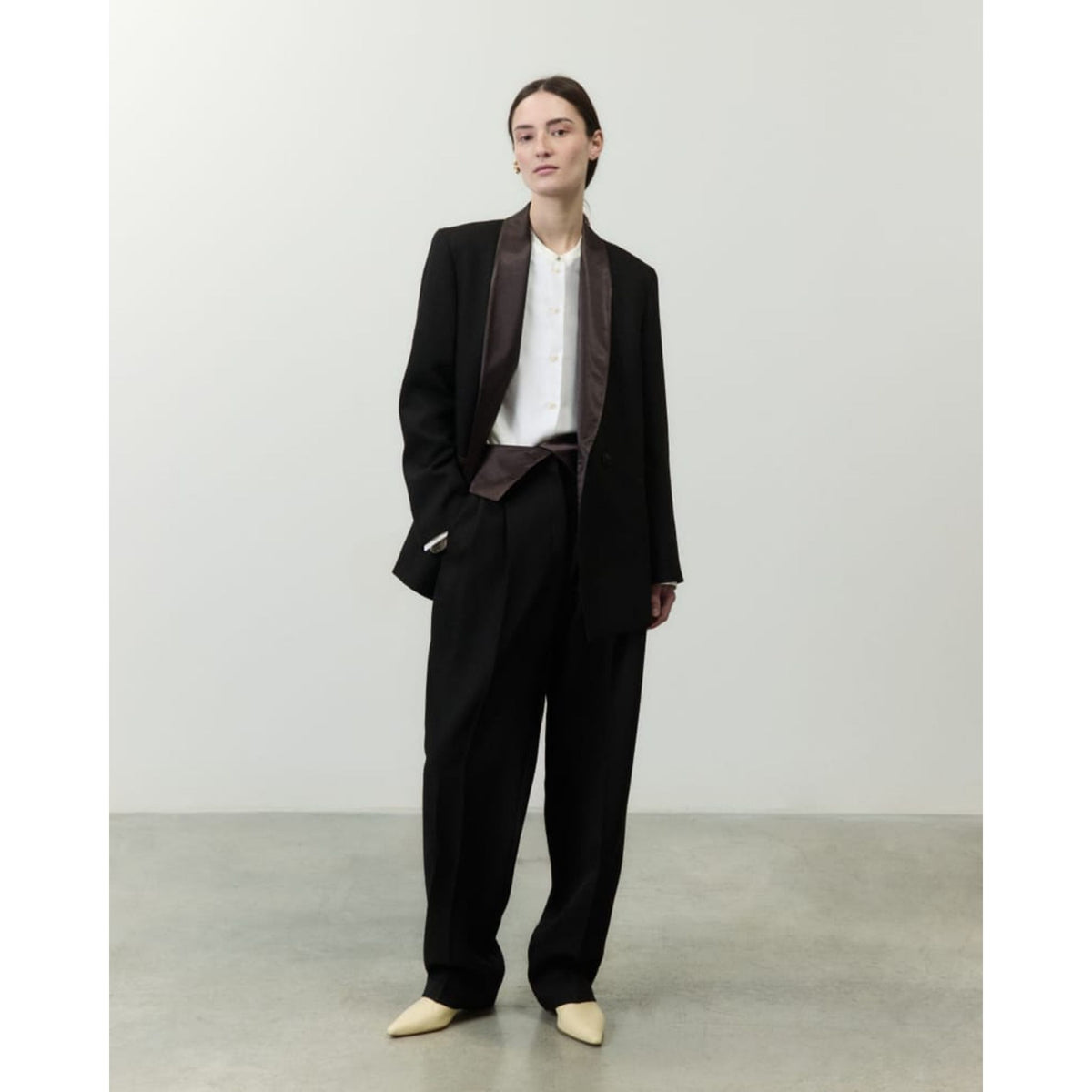 A model stands in a minimalist studio wearing a black wide-leg suit with dark brown satin shawl lapels, a white shirt, and beige pointed-toe shoes.