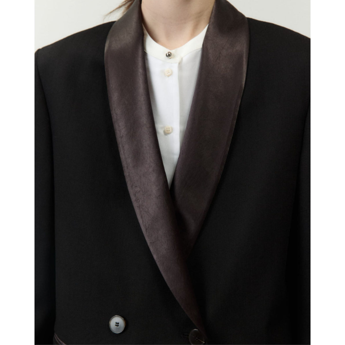 Close-up of a black double-breasted blazer with a wide dark brown satin shawl collar, worn over a white collared shirt.
