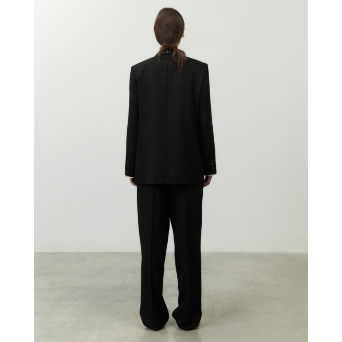A model viewed from the back wearing a black tailored suit with a structured blazer and wide-leg trousers, standing in a minimalist studio setting with a light gray wall and concrete floor.
