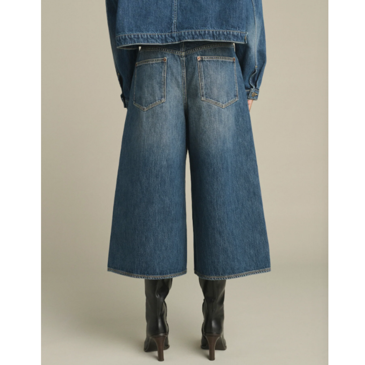 Back view of a model wearing wide-leg blue denim culottes with a faded wash, styled with a denim jacket and black heeled boots against a neutral beige background.