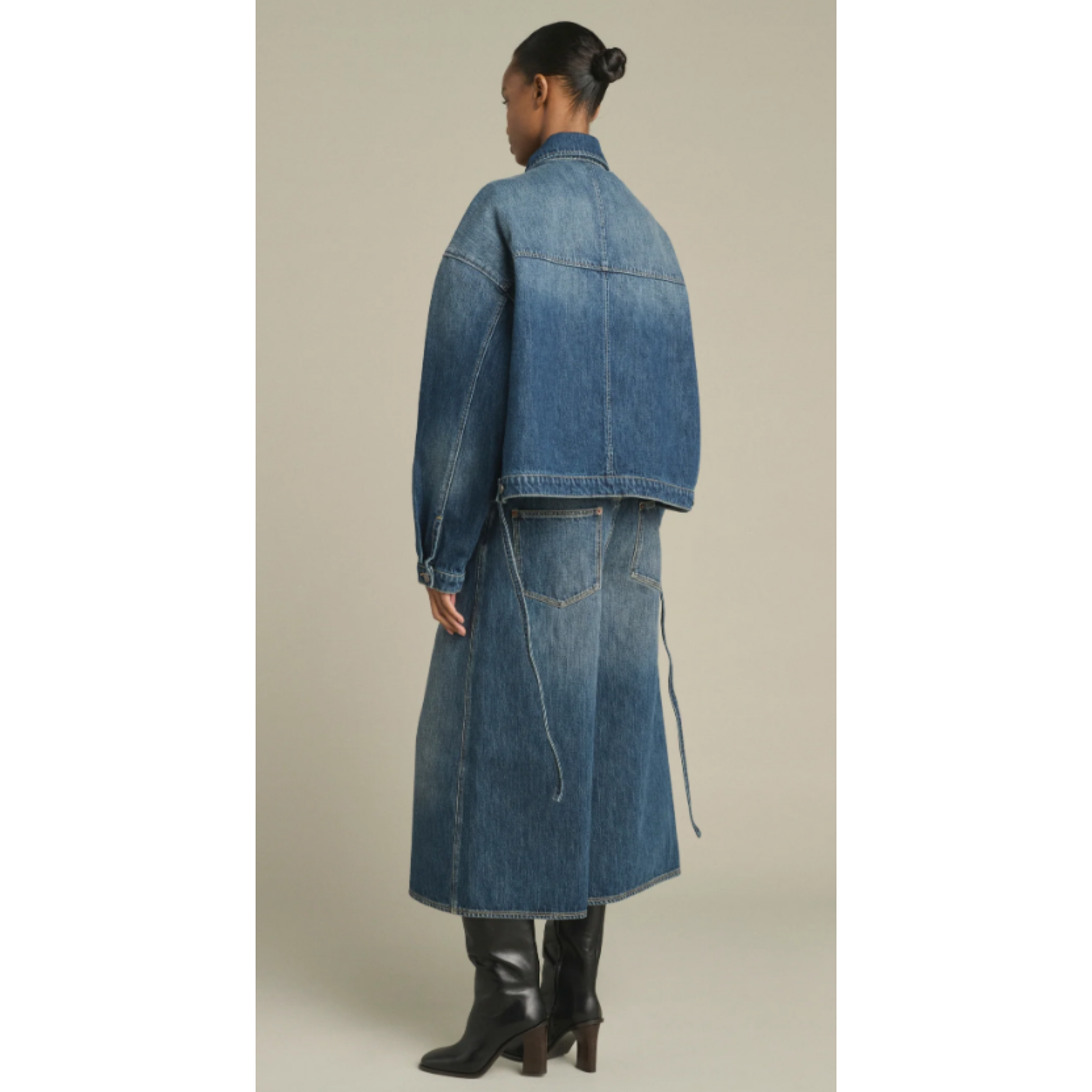 A model wearing a pair of wide-leg cropped denim jeans in medium blue wash, styled with a matching blue denim jacket and black leather ankle boots, set against a plain beige studio background.
