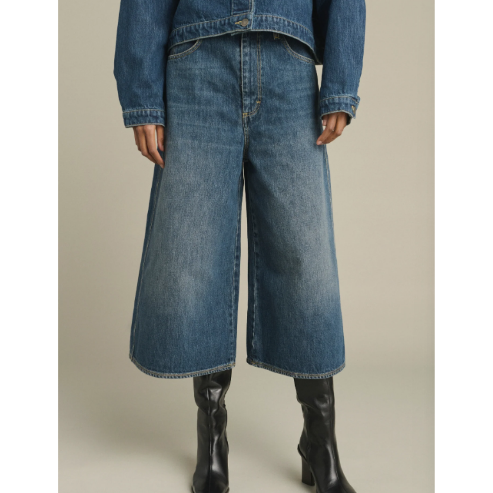 A model wearing a pair of wide-leg cropped denim jeans in medium blue wash, styled with a matching blue denim jacket and black leather ankle boots, set against a plain beige studio background.