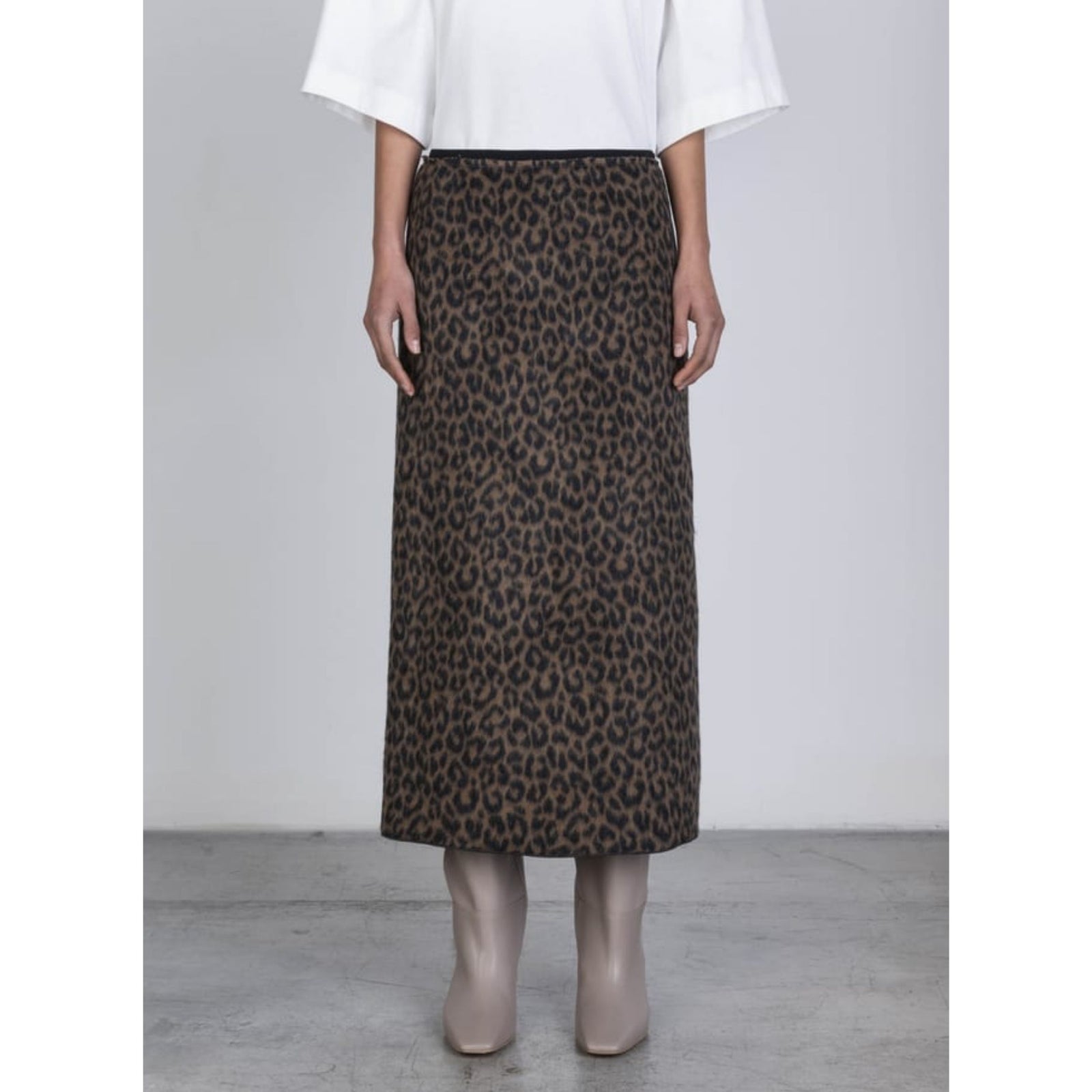 A model wearing a brown and black leopard print midi skirt, paired with a white top and beige boots, standing against a light gray studio background.
