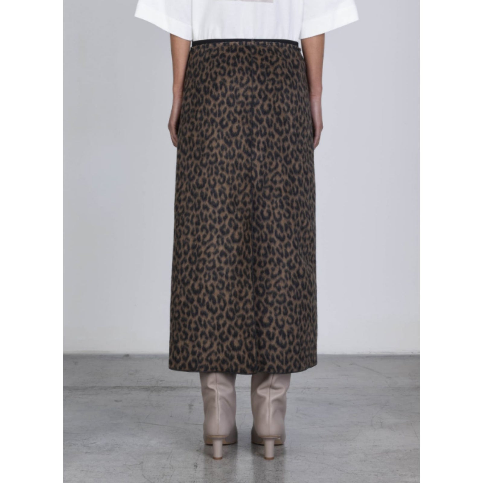 A model wearing a brown and black leopard print midi skirt, paired with a white top and beige boots, standing against a light gray studio background.