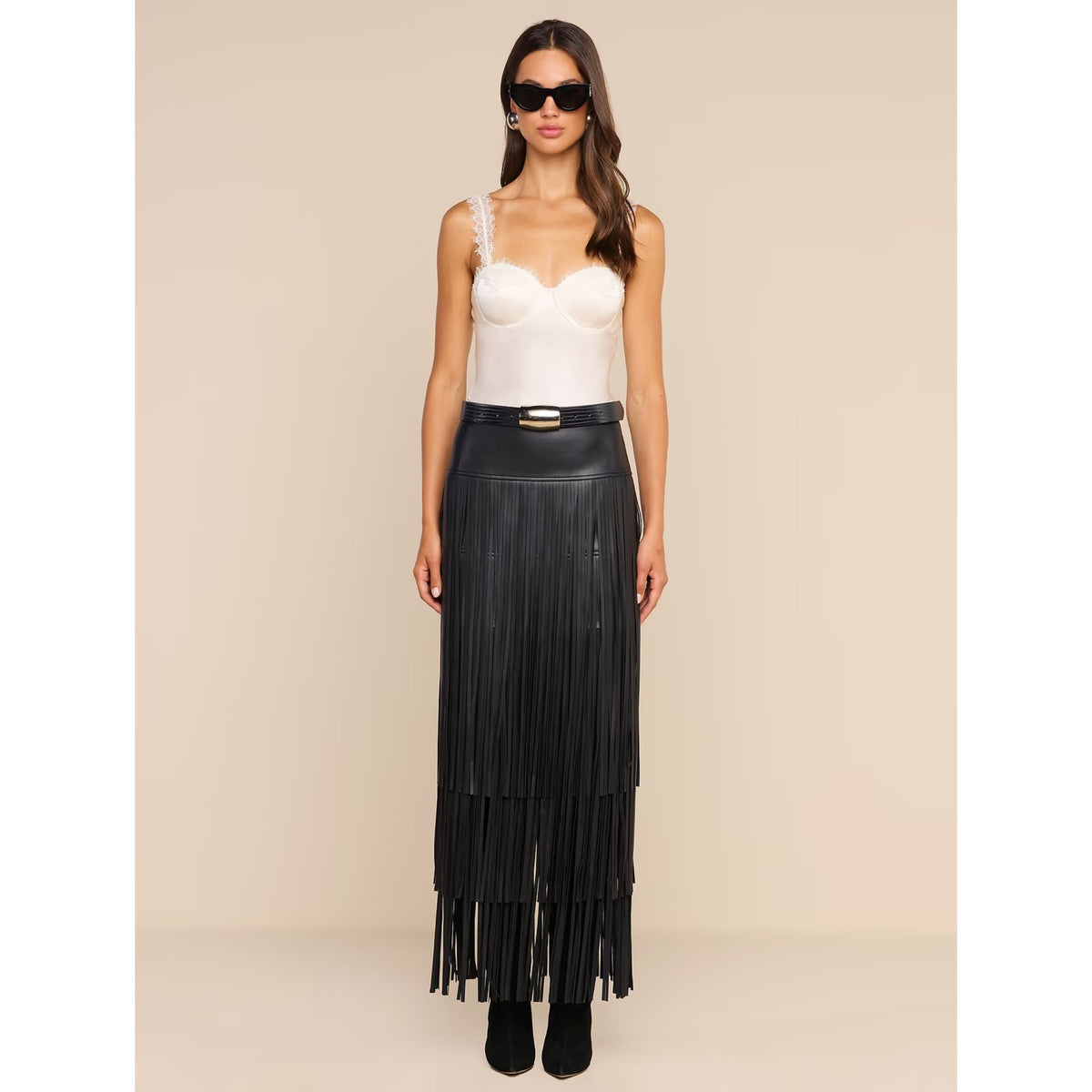 A model stands against a plain beige studio background wearing a white structured camisole top, a black wide belt with a gold buckle, and a long black pleated skirt with multiple horizontal fringe layers. She is also wearing black sunglasses and black ankle boots.