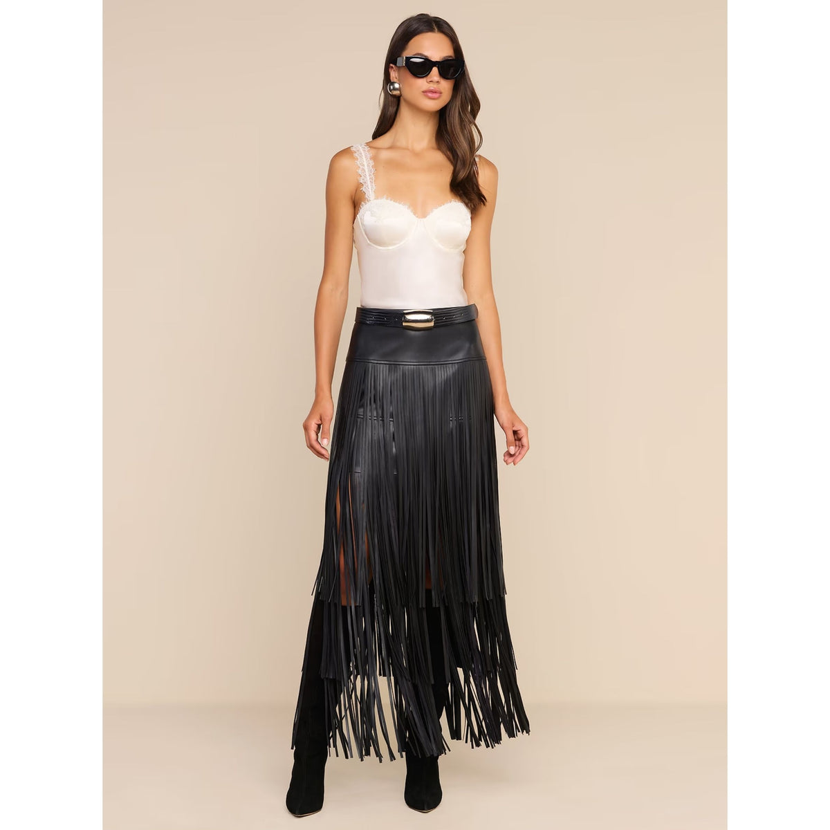A model wearing a long black faux-leather fringe skirt with a gold-tone belt, paired with a white corset-style camisole and black boots, set against a solid beige studio background.