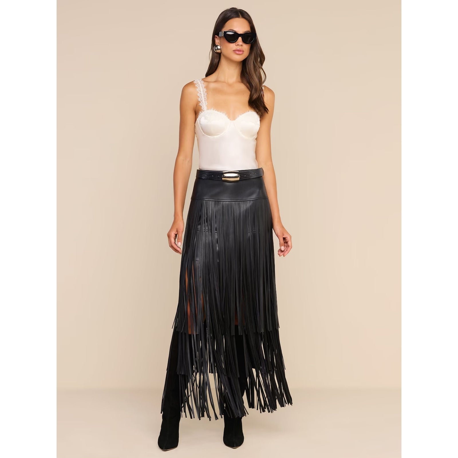 A model wearing a long black faux-leather fringe skirt with a gold-tone belt, paired with a white corset-style camisole and black boots, set against a solid beige studio background.