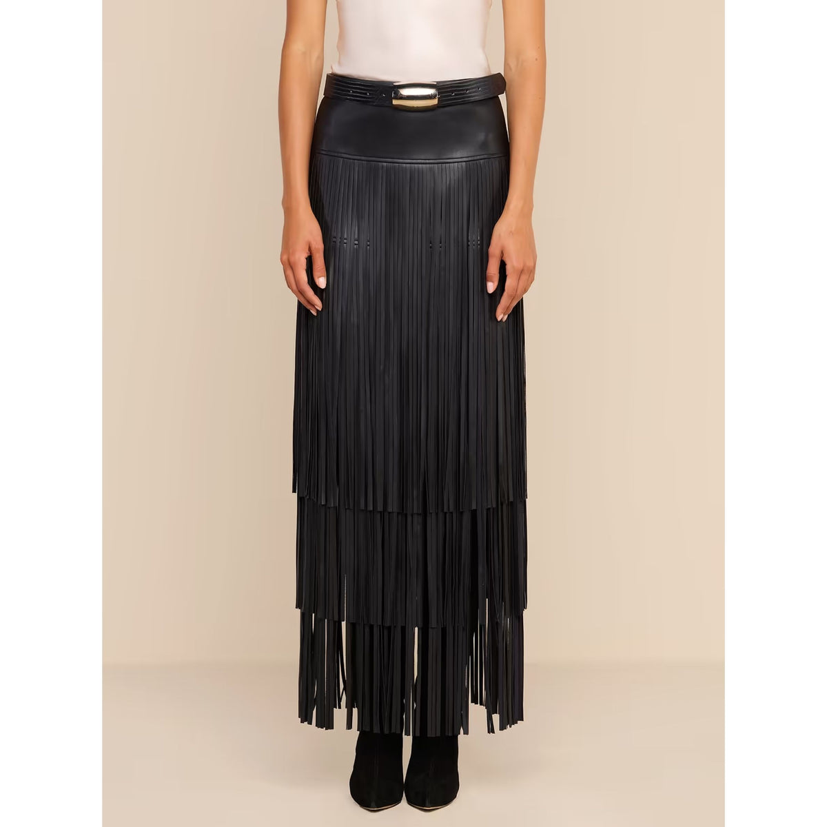 A model wearing a long black faux-leather pleated maxi skirt with a matching belt featuring a gold buckle, styled with a white top and black ankle boots against a beige studio background.