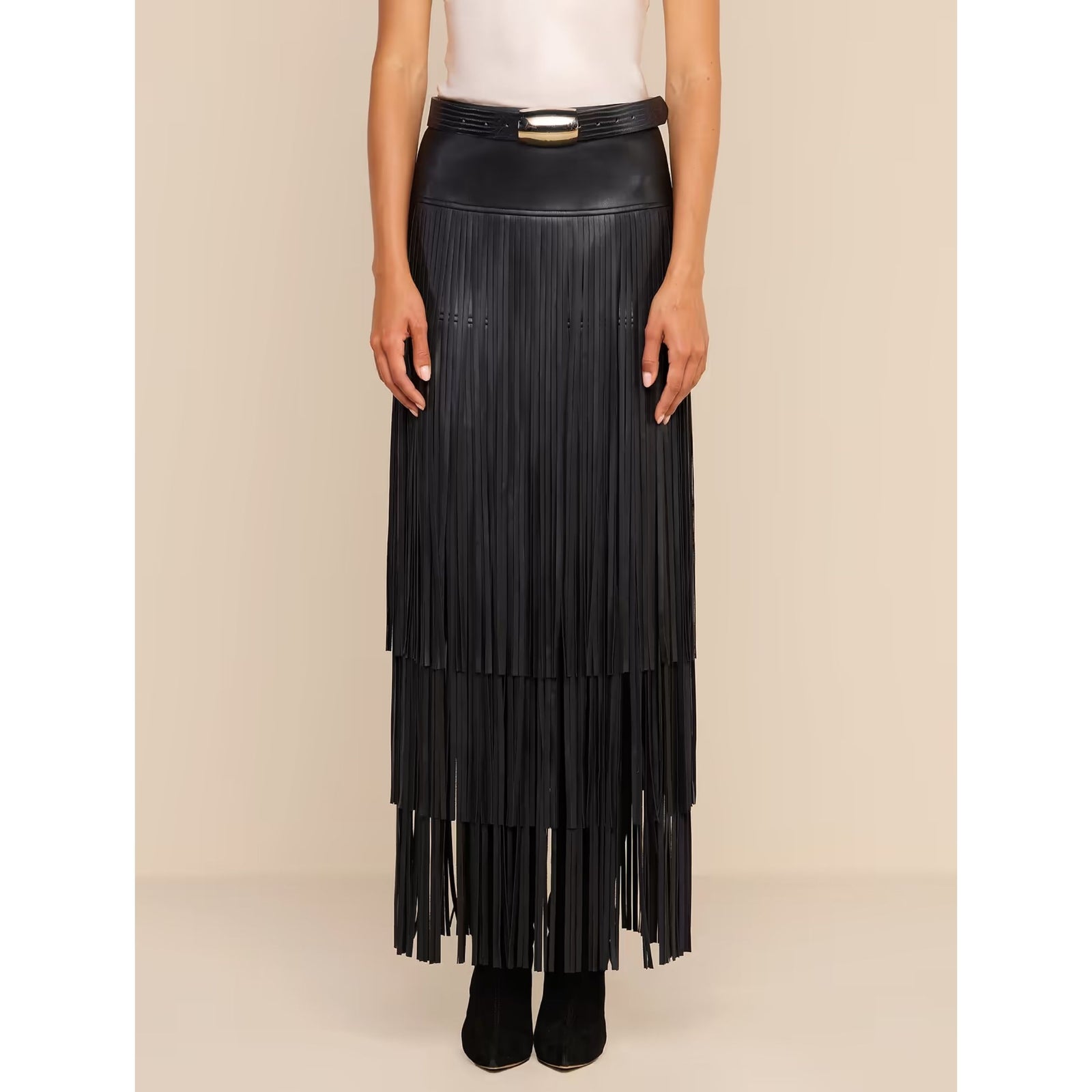 A model wearing a long black faux-leather fringe skirt with a gold-tone belt, paired with a white corset-style camisole and black boots, set against a solid beige studio background.