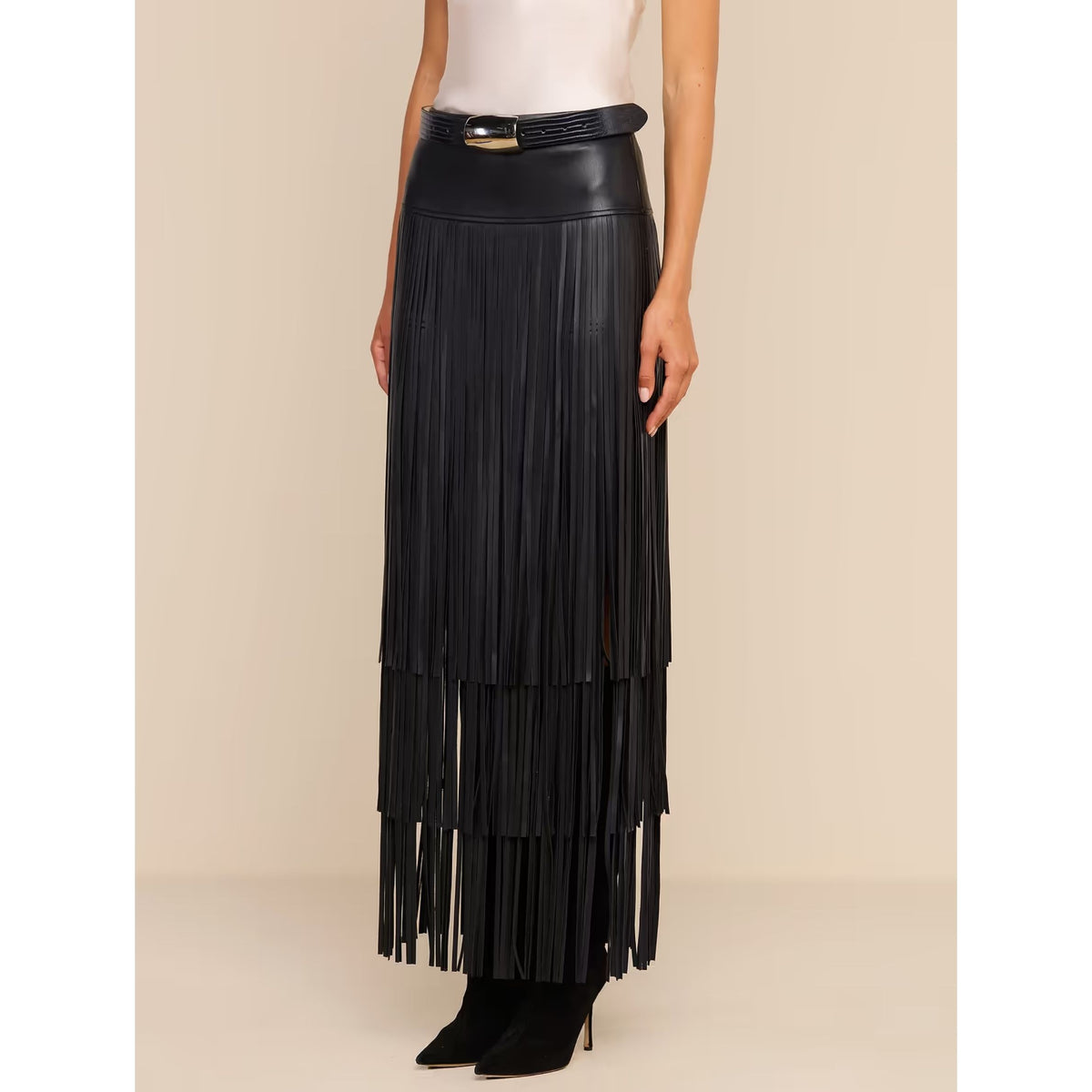 A black faux-leather fringe maxi skirt with a matching belt and gold buckle, shown on a model against a beige studio background.