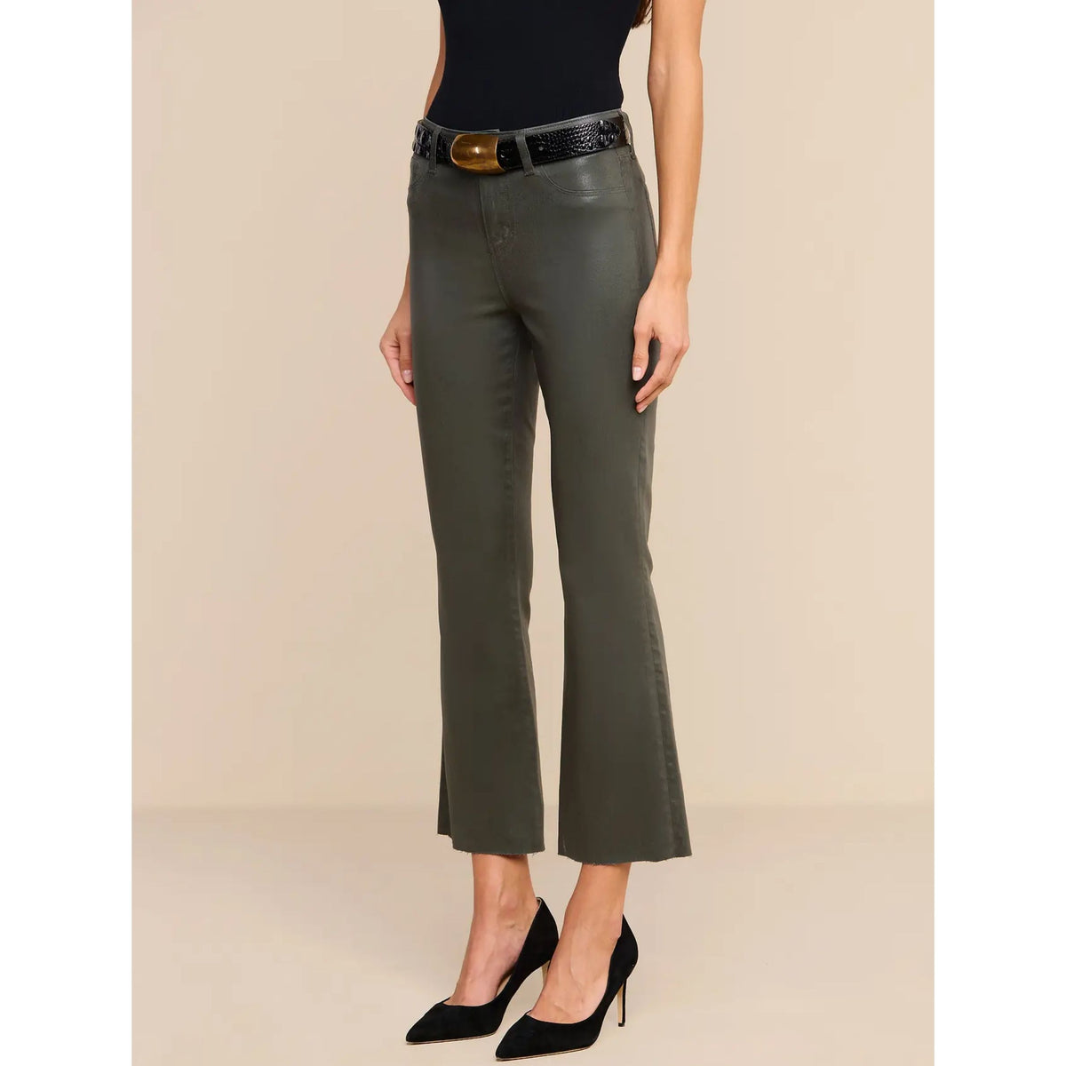 A model wearing dark olive green faux-leather cropped flare pants with a black belt and gold buckle, styled with a black bodysuit and black pointed-toe heels against a beige studio background.