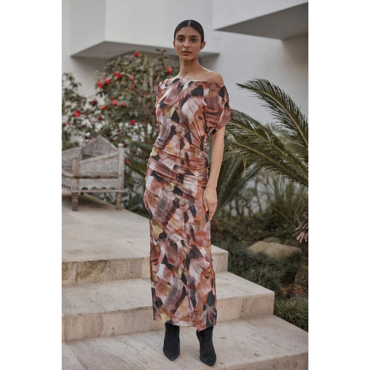 A woman models an abstract print midi dress with a dusty rose, brown, and black pattern, styled with black ankle boots in an outdoor patio setting with stone steps and greenery.