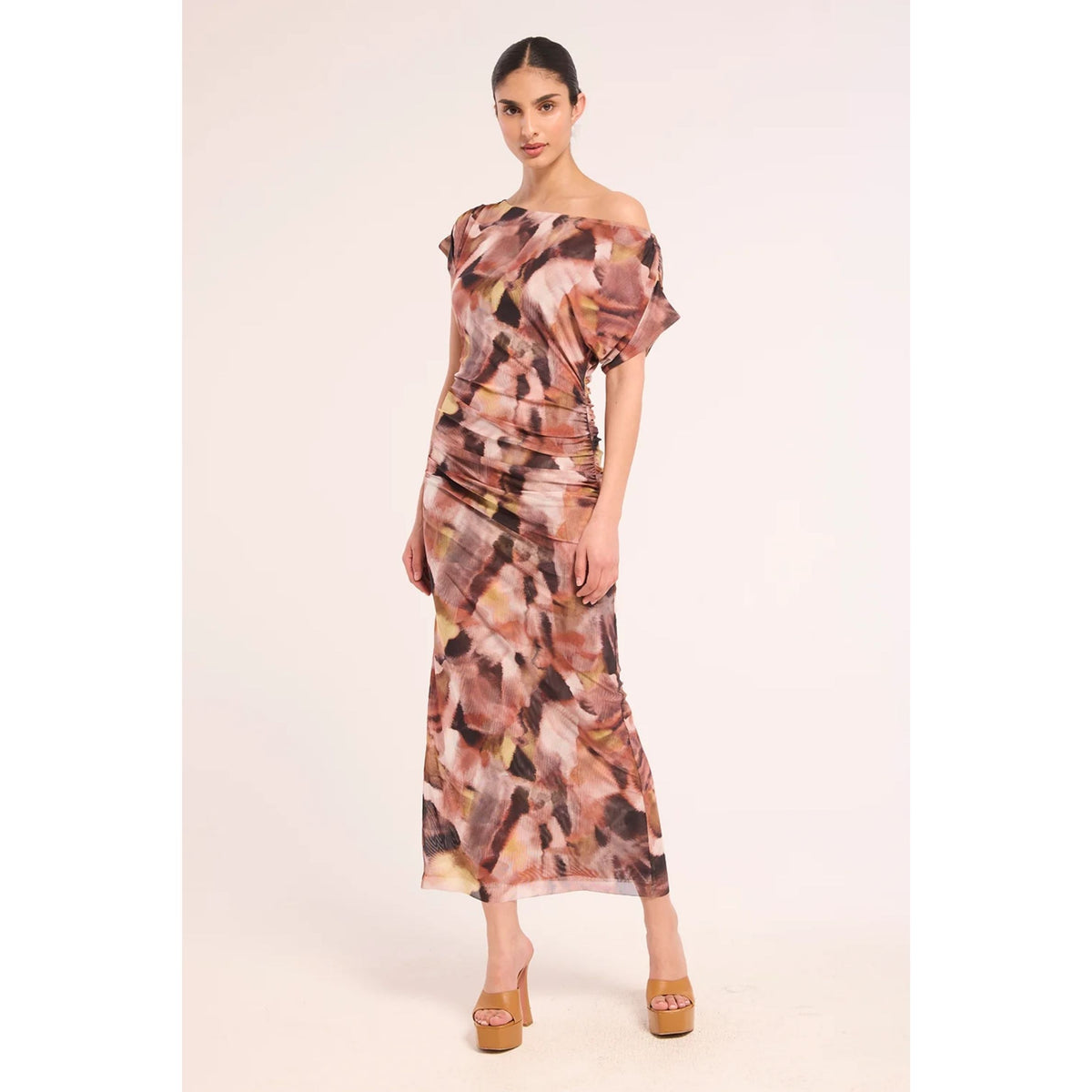 A woman modeling a draped off-the-shoulder midi dress with an abstract painterly pattern in shades of brown, beige, and black, paired with tan platform sandals against a solid light beige studio background.