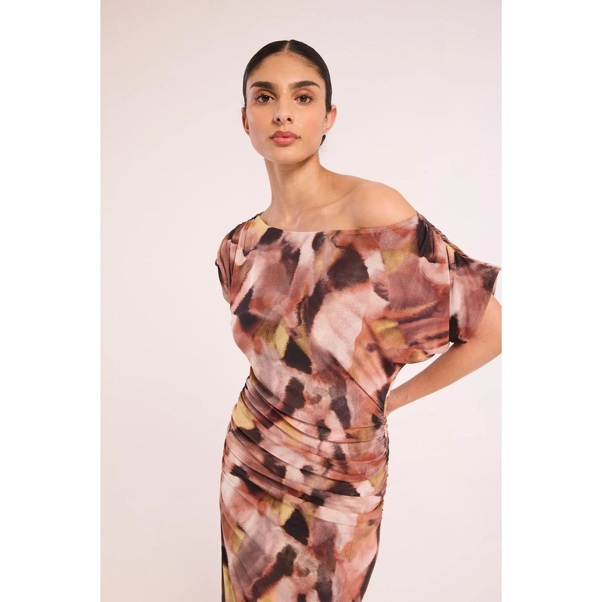 A woman modeling an off-the-shoulder, ruched midi dress with an abstract watercolor print in dusty pink, brown, beige, and olive green tones, set against a plain light beige studio background.