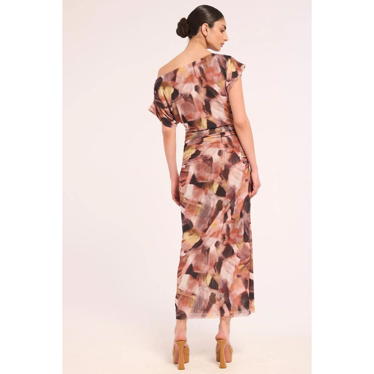 A woman modeling a one-shoulder midi dress with a multicolor abstract print in pink, brown, beige, and black, shown from a back-three-quarter angle against a light pink studio background.