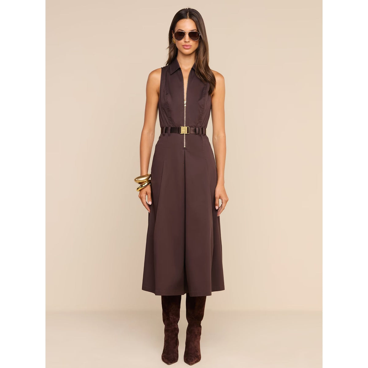 A woman modeling a sleeveless dark brown midi dress with a front zipper, a matching dark brown belt with a gold-tone buckle, brown suede boots, and sunglasses against a solid beige studio background.
