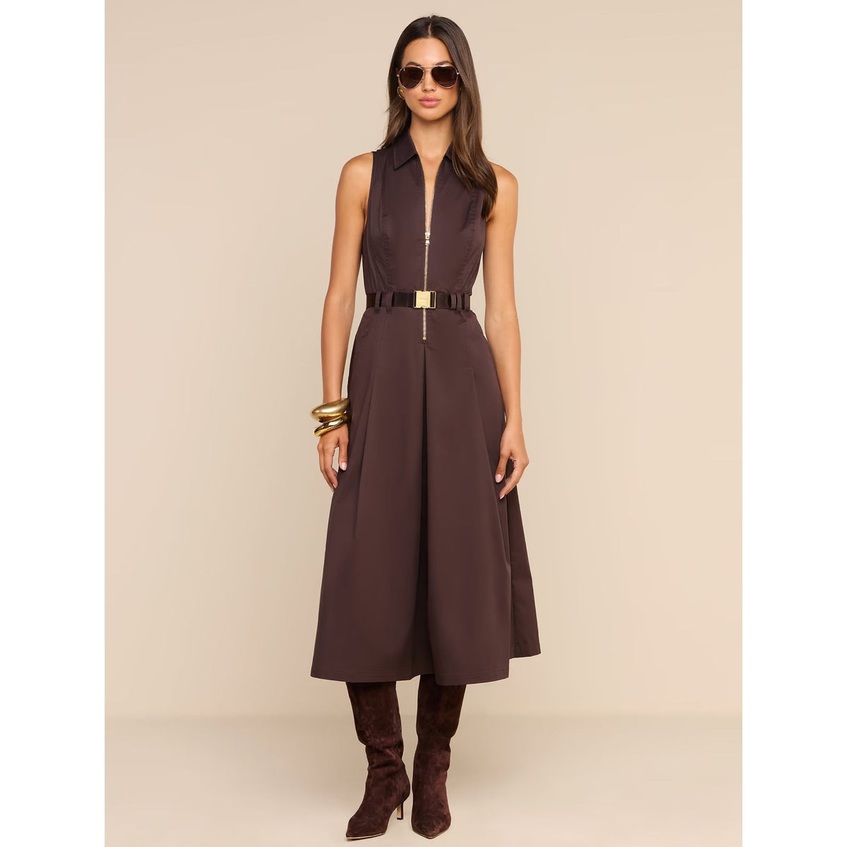 A woman models a sleeveless dark brown midi dress with a front zipper, a matching dark belt with a gold-tone buckle, sunglasses, gold bracelets, and dark brown suede boots against a solid beige studio background.