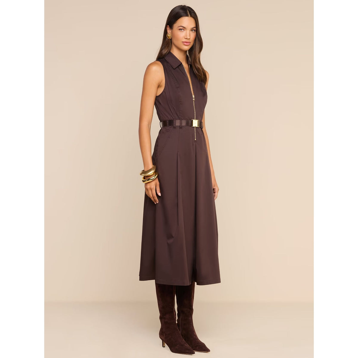 A woman models a sleeveless dark brown midi dress with a collar, a front zipper, a matching dark belt with a gold buckle, and dark brown heeled boots against a solid beige studio background.