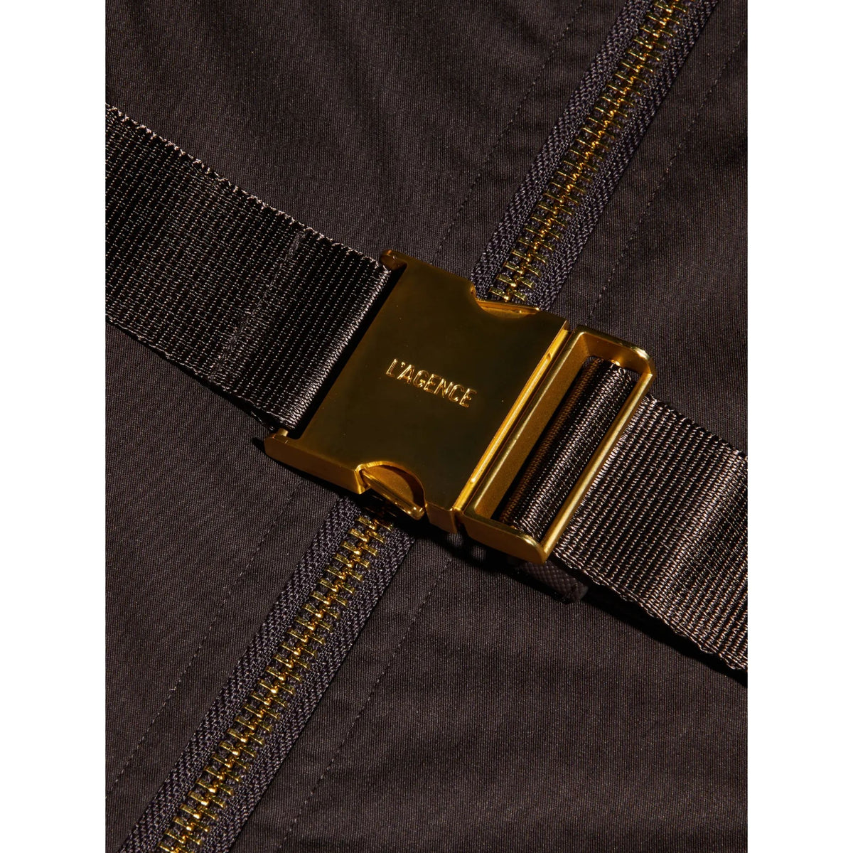 Close-up of a dark brown or black nylon garment with a prominent gold-tone metal buckle and zipper, featuring the embossed brand name 'LYENCE' on the buckle.