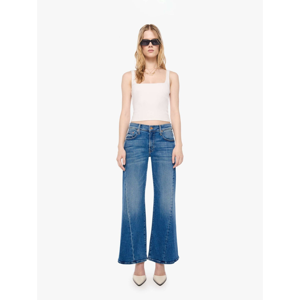 A model wearing blue wide-leg flare jeans, a cream sleeveless crop top, and beige pointed-toe heels, standing against a plain white studio background.