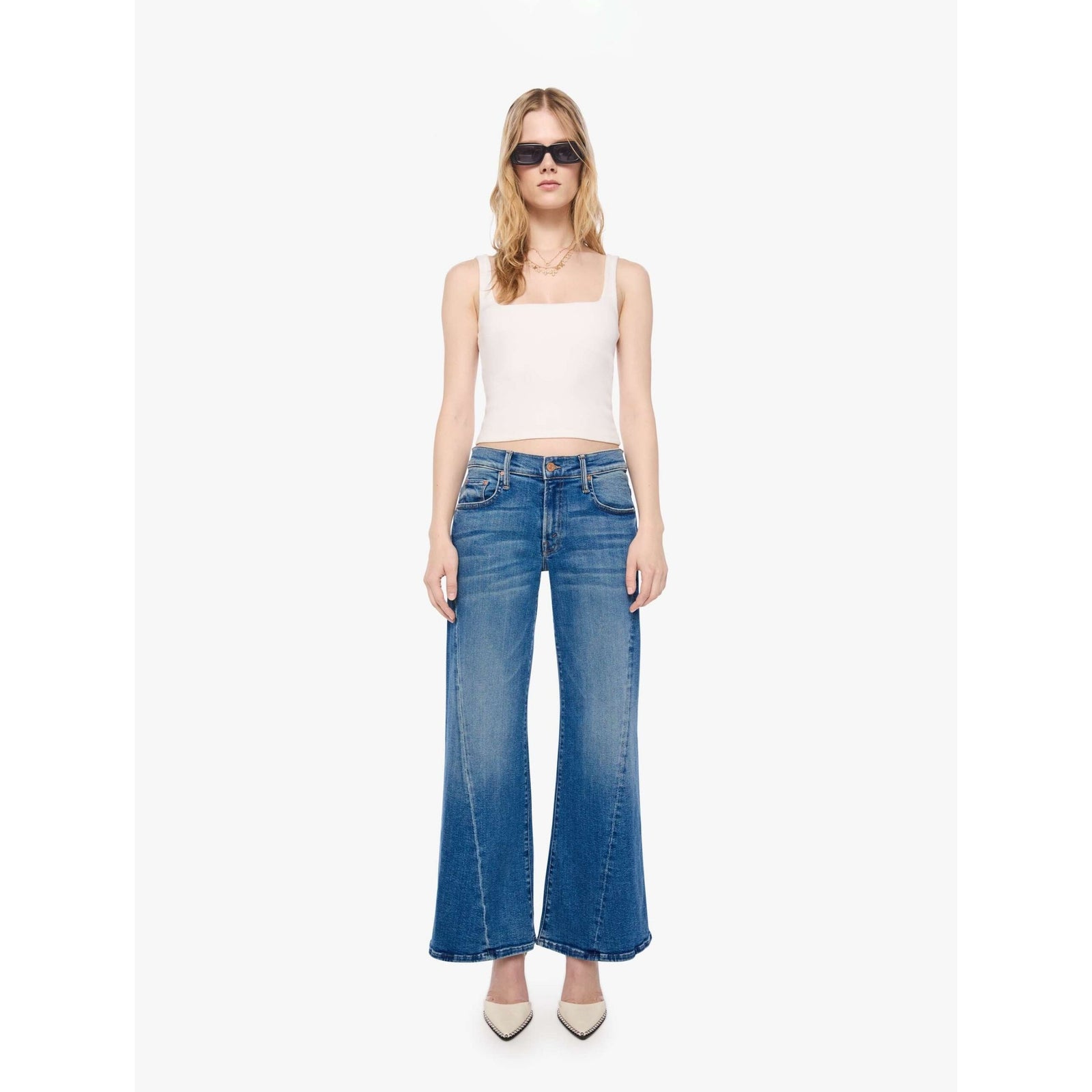 A model wearing blue wide-leg flare jeans, a cream sleeveless crop top, and beige pointed-toe heels, standing against a plain white studio background.